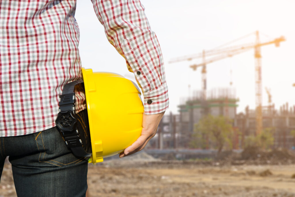 Hand's engineer worker holding yellow safety helmet with building on site background.